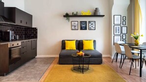 Eine moderne Küche mit dunklen Schränken, einer schwarzen Arbeitsplatte und einem gelben Teppich. Es gibt eine Essecke mit Tisch und Stühlen sowie eine Couch mit gelben Kissen.