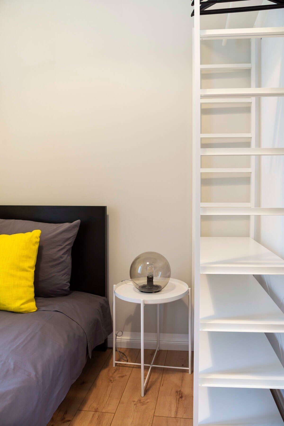 A bedroom with a bed, a bedside table with a lamp, and a white shelving unit.