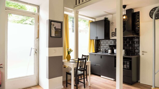 A modern kitchen with black cabinets, a dining area with a white table and chairs, and a view of the outdoors through a door. - Charmante Wohnung in Berlin-Wilmersdorf (Berlin)