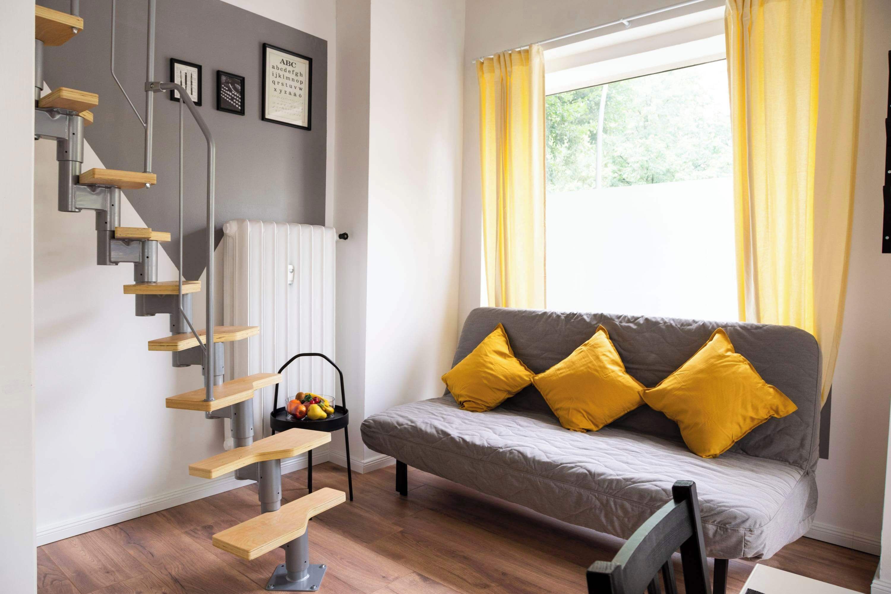 Una sala de estar moderna con un sofá gris, cojines amarillos, una escalera de madera y una ventana con cortinas amarillas.