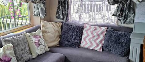 A cozy corner with a grey sectional sofa, patterned cushions, a black rug, and a window with patterned curtains.