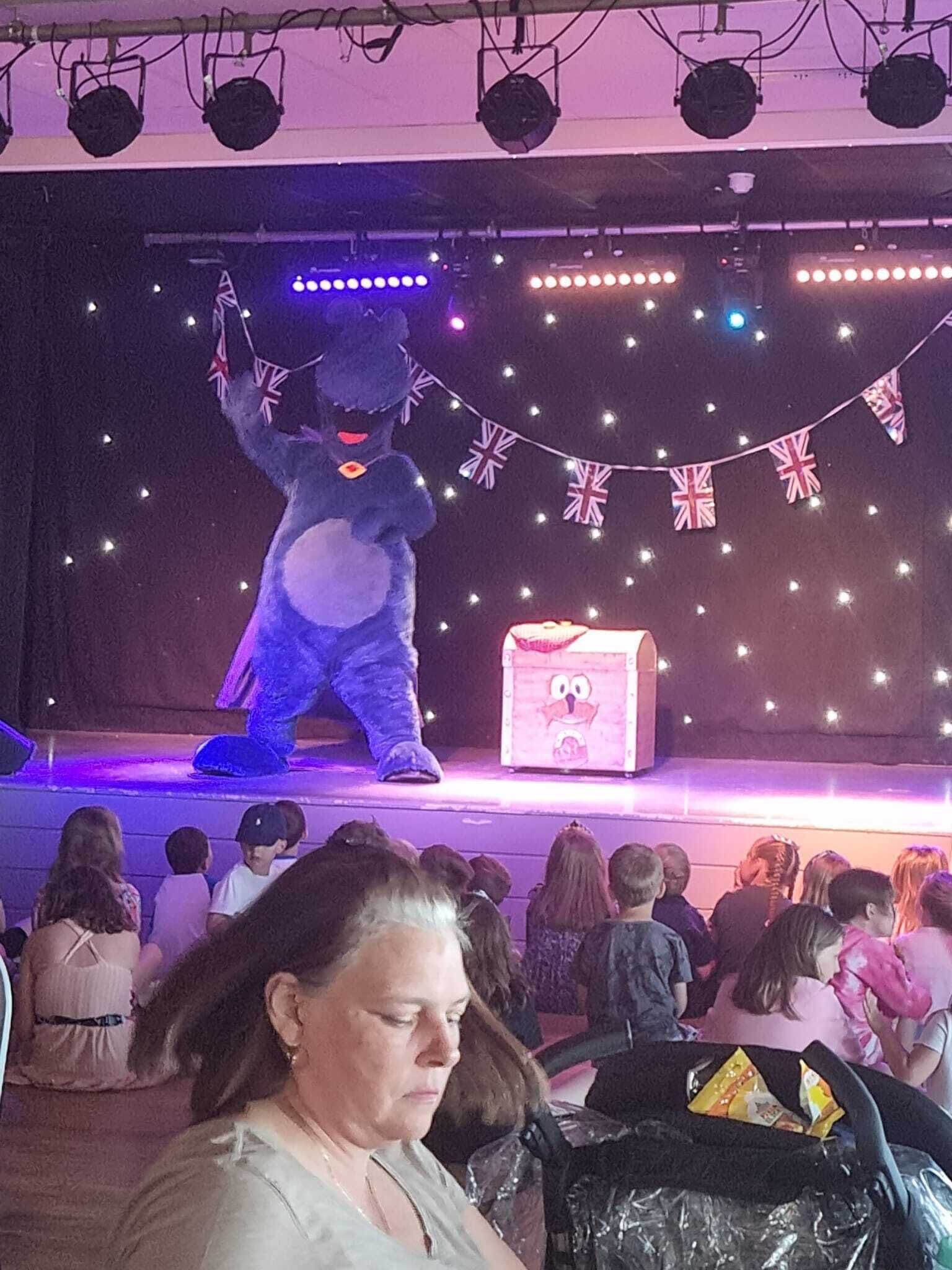 A stage with a character in a blue costume, a pink box with a face, and string lights.