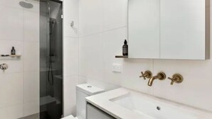 A modern bathroom with a glass shower enclosure, a white sink, and a mirror with a gold faucet. - New Spacious 2 Bedroom With Gorgeous City Views (Northcote)