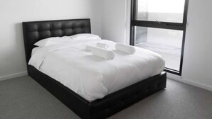 A modern hotel room with a black headboard, white bedding, and a large window offering a view of the cityscape. - New Spacious 2 Bedroom With Gorgeous City Views (Northcote)