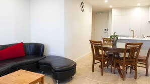A living room with a black leather sofa, a red pillow, a wooden coffee table, and a dining area with wooden chairs and a glass table. - New Spacious 2 Bedroom With Gorgeous City Views (Northcote)