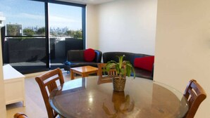 Een moderne woonkamer met een glazen tafel, houten stoelen, een bank met rode kussens en een groot raam met uitzicht op de stad.