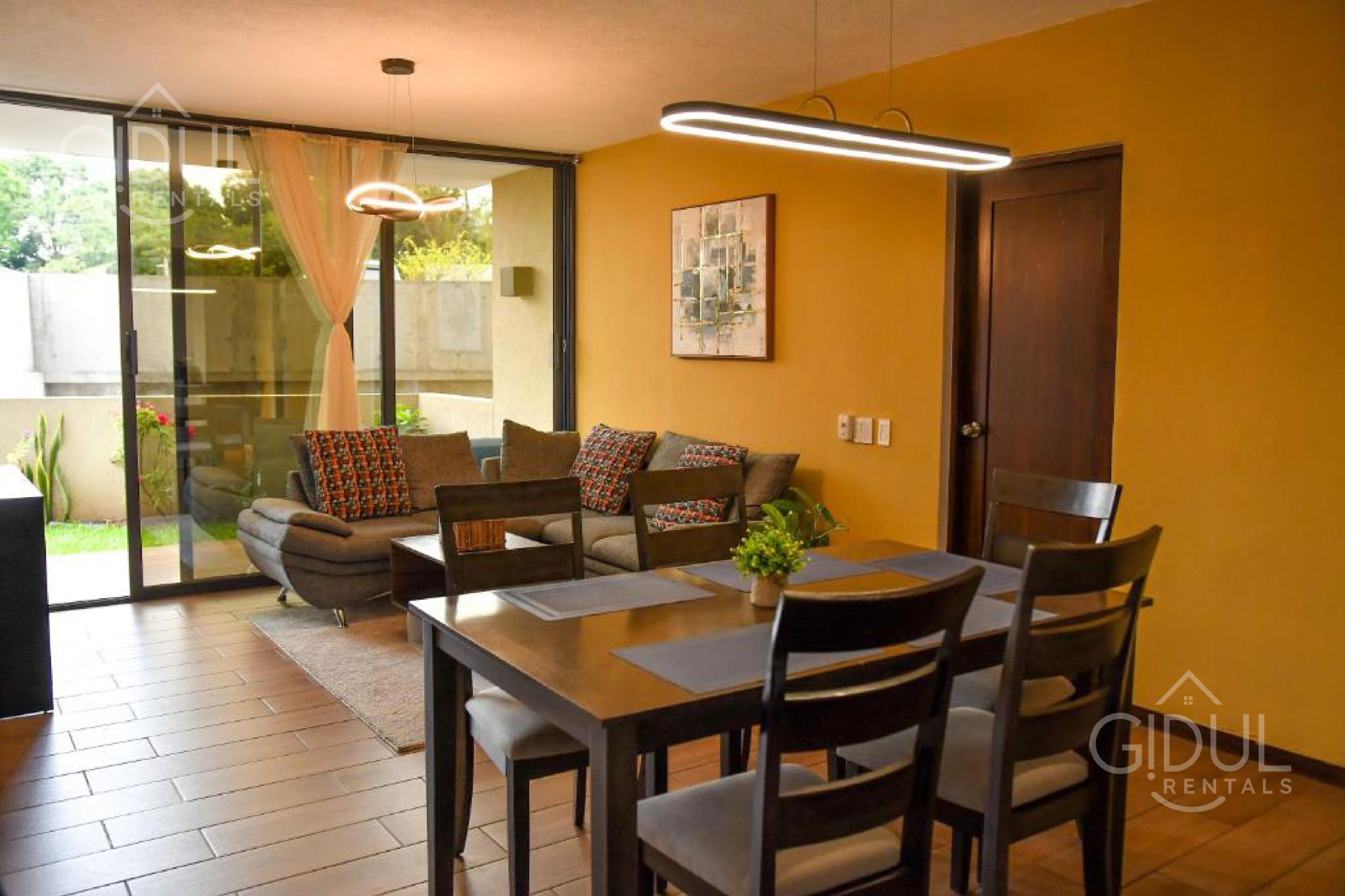 A modern living room with a sofa, dining table, and chairs. There is a sliding glass door leading to a balcony.