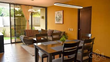 A modern living room with a sofa, dining table, and chairs. There is a sliding glass door leading to a balcony.