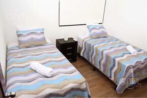 Quarto com duas camas, cada uma com roupa de cama e almofadas listradas, uma mesa de cabeceira com abajur e um quadro na parede.