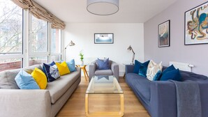 Una sala moderna con un sofá gris, un sofá azul y una mesa de centro de vidrio. Hay varios cojines en amarillo y azul, y una lámpara sobre una mesita auxiliar.