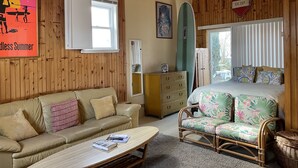 Tunnelmallinen huone, jossa on puinen katto, beigenvärinen sohva, rottinkituoli, kukallisilla tekstiileillä varustettu sänky ja seinällä surffilauta.