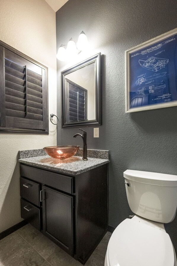 A bathroom with a copper vessel sink, dark cabinetry, a white toilet, and a wall-mounted mirror.