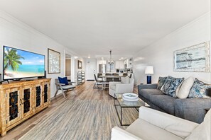A modern living room with a television, a sofa, a coffee table, and a dining area with a chandelier. - Huntington By the Sea 306. Gulf Front, Community Pool, Free Beach Service. (Miramar Beach)