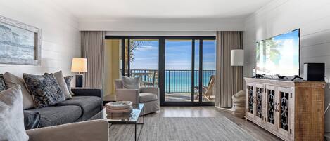A modern living room with a sofa, armchairs, a coffee table, and a TV on a wooden cabinet. A large window offers a view of the ocean.