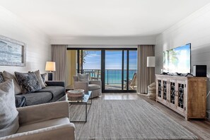 A modern living room with a sofa, armchairs, a coffee table, and a TV on a wooden cabinet. A large window offers a view of the ocean.
