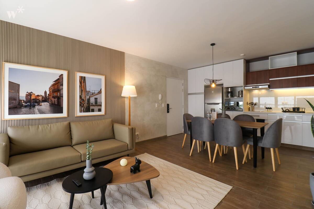 Ein modernes Wohnzimmer mit einer Couch, Couchtisch und Essbereich mit Stühlen. An der Wand befinden sich gerahmte Bilder, eine Lampe und eine Küchenzeile mit Schränken und Geräten.