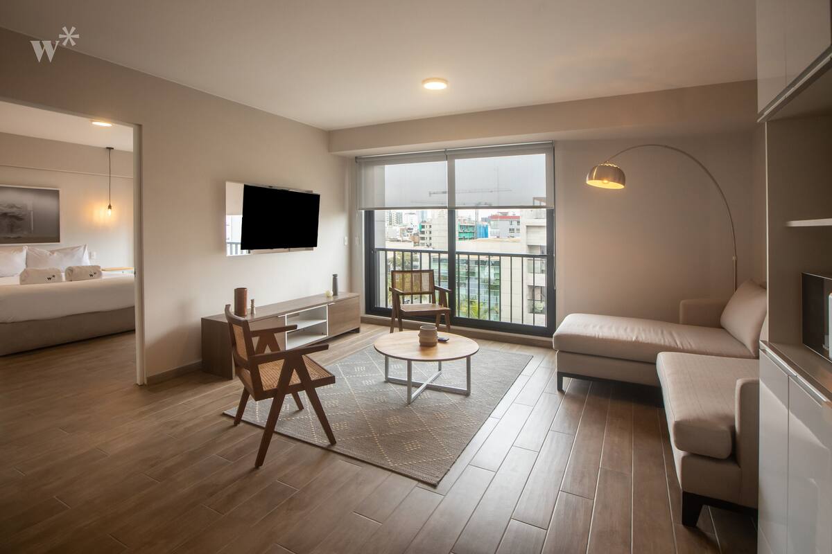Ein modernes Wohnzimmer mit einer Couch, einem Esstisch mit Stühlen, einem an der Wand befestigten Fernseher und einem großen Fenster mit Blick auf Gebäude.