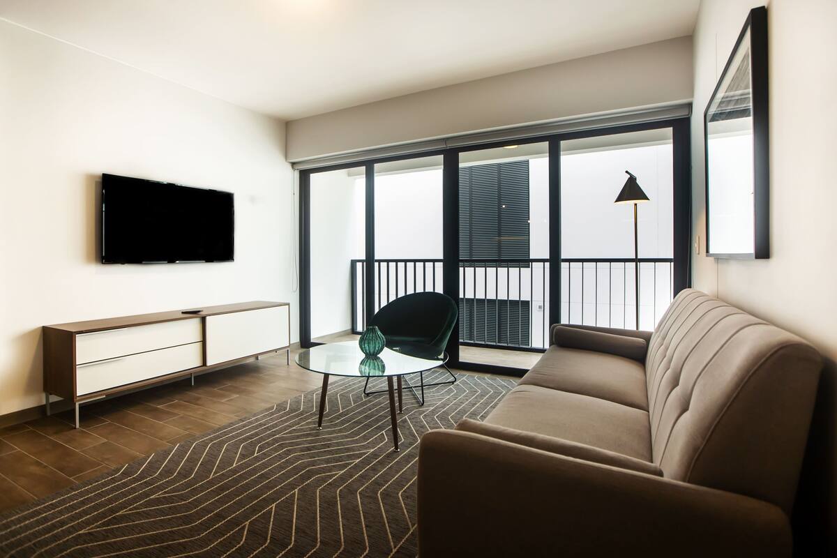 Ein modernes Wohnzimmer mit einer Couch, einem Glastisch, einem an der Wand befestigten Fernseher und einem Balkon mit Blick.