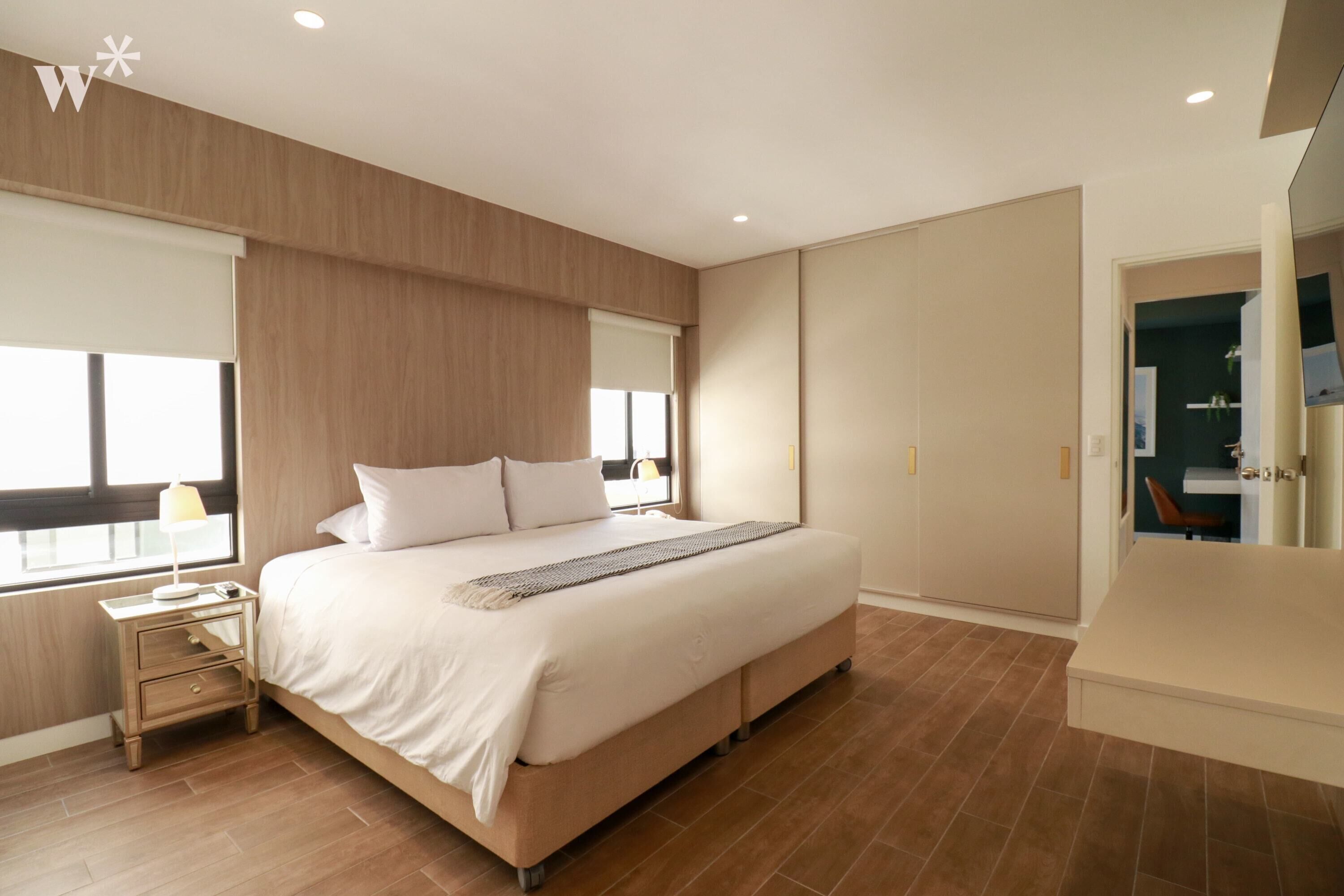 A modern bedroom with a large bed, wooden panel walls, a nightstand, and a view of a bathroom.