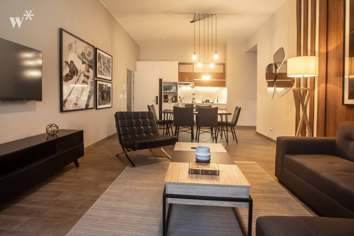 Ein modernes Wohnzimmer mit Flachbildfernseher, schwarzem Ledersofa und einem Essbereich mit Holztisch und Stühlen.