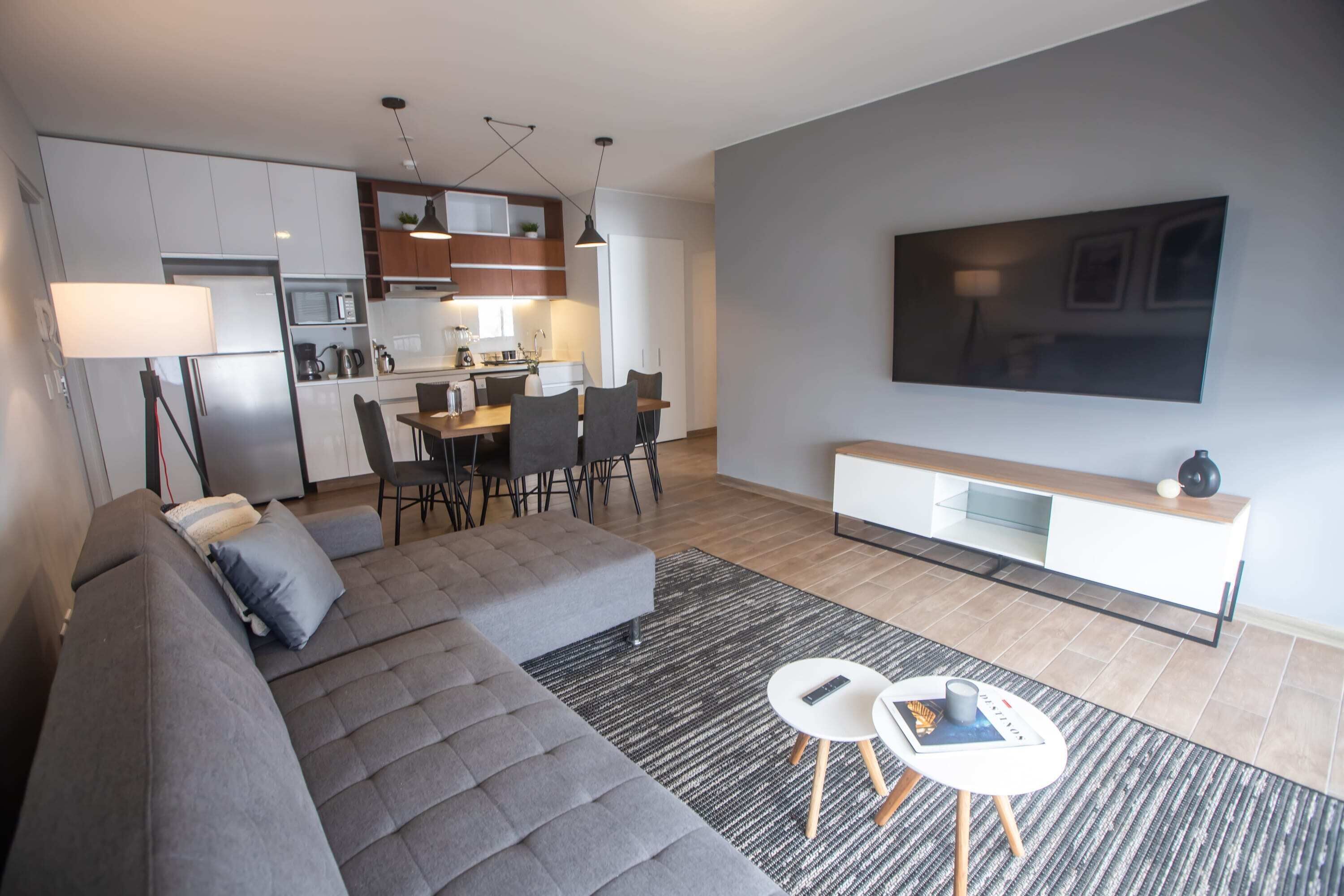 A modern living room with a grey sectional sofa, a dining area with a table and chairs, a kitchen with white cabinets, and a large flat-screen TV mounted on the wall.