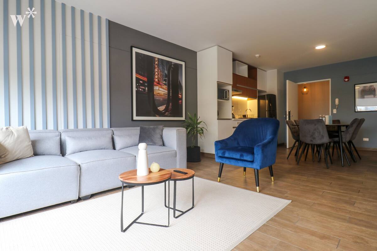 Ein modernes Wohnzimmer mit grauem Sofa, blauem Sessel und einem Essbereich mit Tisch und Stühlen.