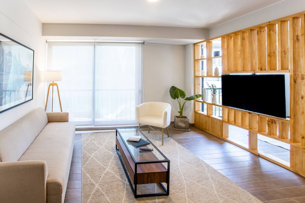 Ein modernes Wohnzimmer mit einer Couch, einem Glastisch, einem weißen Sessel, einer Holz-TV-Konsole und einem großen Fernseher.