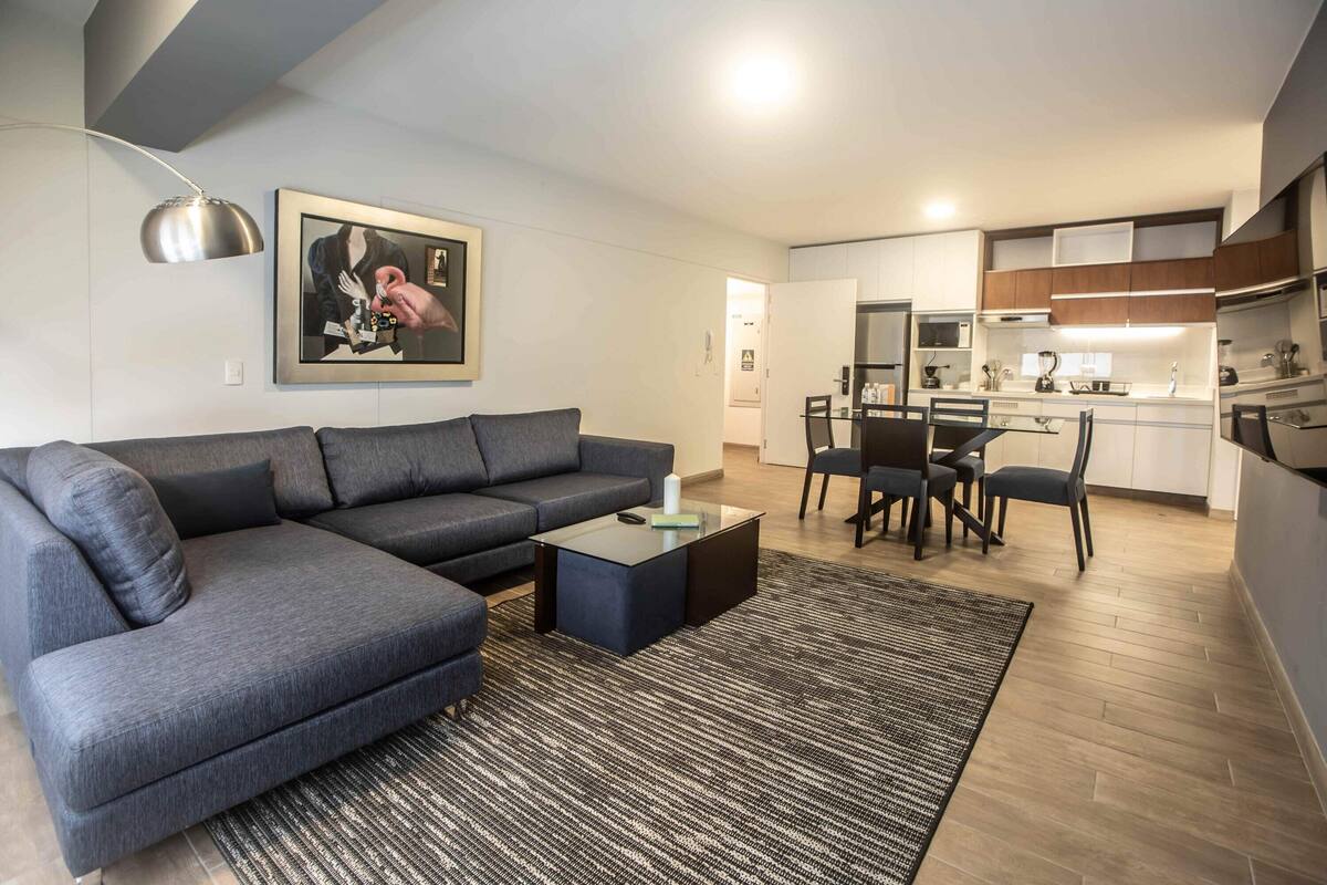Ein modernes Wohnzimmer mit einem grauen Ecksofa, einem Glastisch und einem Essbereich mit Tisch und Stühlen.