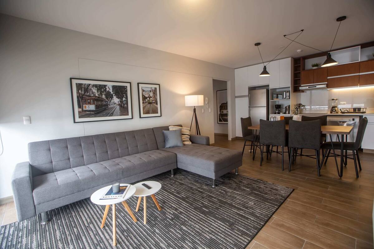 Ein modernes Wohnzimmer mit einem grauen Ecksofa, einem kleinen runden Couchtisch und einem Essbereich mit Holztisch und Stühlen.