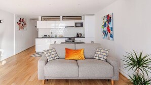 Ein modernes Wohnzimmer mit grauem Sofa, Holzboden, einer Küche im Hintergrund und einem großen abstrakten Gemälde an der Wand.