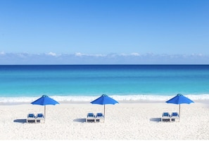 Una playa con tres sombrillas azules y dos sillas reclinables blancas.