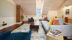A loft-style living room with a high ceiling, exposed wooden beams, a brick wall, a blue sofa, a grey sofa, a wooden coffee table, and a view through a skylight. - ★Farmhouse Style★Outdoor Patios★Parking!★Beautiful (Pittsburgh)