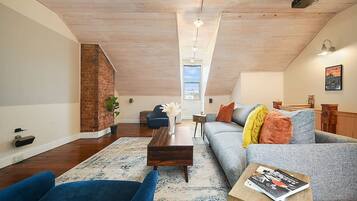 A loft-style living room with a high ceiling, exposed wooden beams, a brick wall, a blue sofa, a grey sofa, a wooden coffee table, and a view through a skylight.