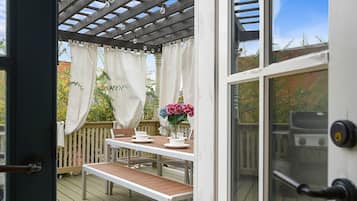 An outdoor deck with a wooden table and chairs, a pergola with white curtains, and a view of the surrounding garden.