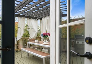 An outdoor deck with a wooden table and chairs, a pergola with white curtains, and a view of the surrounding garden.