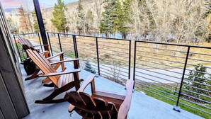 A covered outdoor area with wooden chairs and a metal railing, overlooking a forested landscape. - Brand new property with hot tub, fire pit, parking, covered ski storage, and more! (Silverthorne)