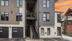 A modern multi-story building with a combination of wood and metal siding, a central staircase, and a garage with two doors. - Brand new property with hot tub, fire pit, parking, covered ski storage, and more! (Silverthorne)