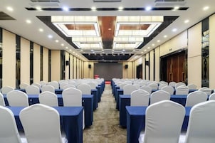 Un salón de conferencias con filas de mesas y sillas, un escenario al frente y un techo alto con tragaluces.