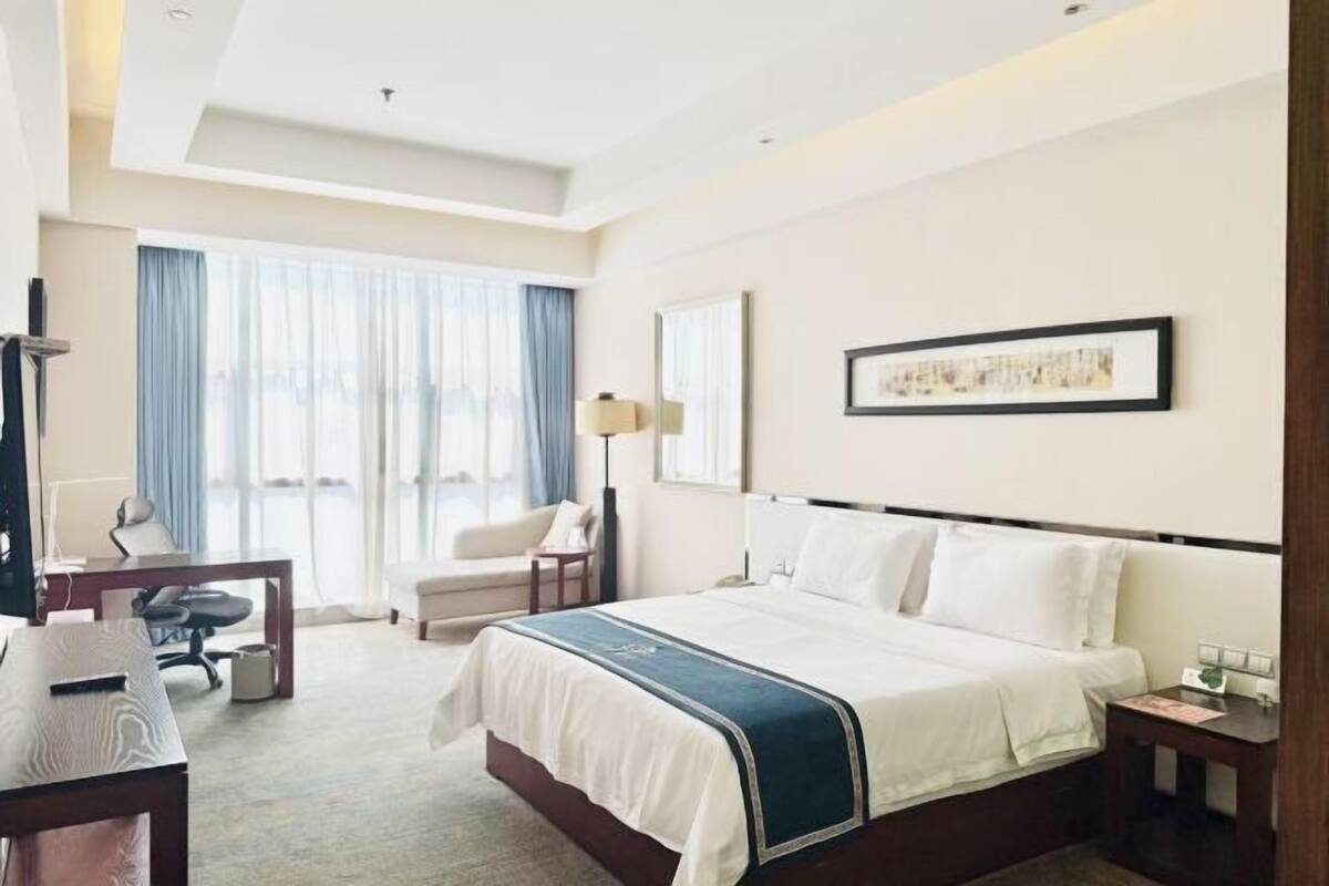 A hotel room with a large bed, a desk, a chair, a lamp, and a window with curtains.