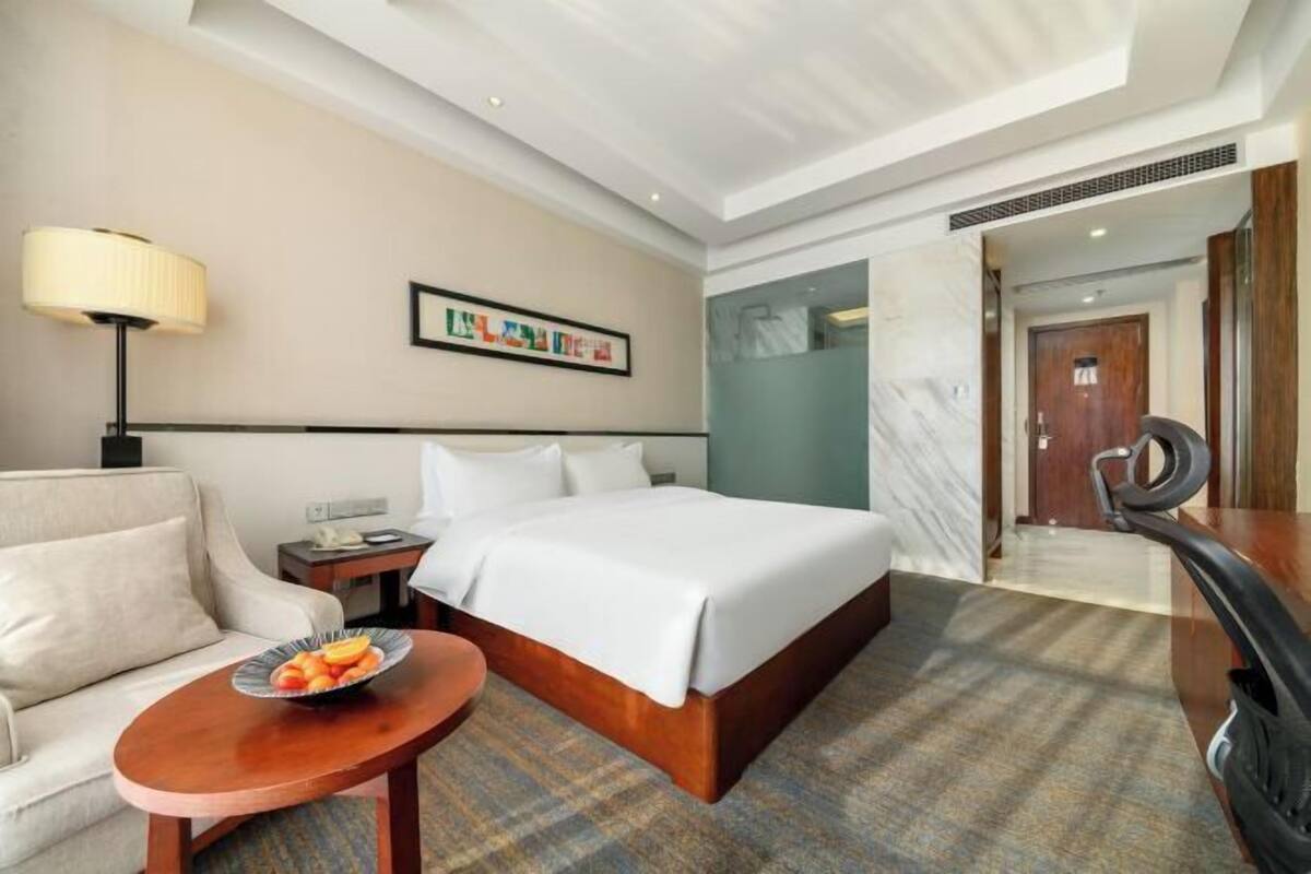 A hotel room with a bed, a sofa, a small table with fruit, a lamp, and a framed picture on the wall.