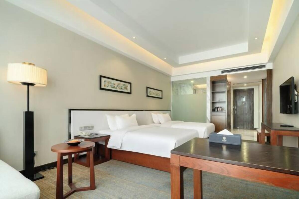 A hotel room with two beds, a desk, a chair, a lamp, a TV, and a minibar.
