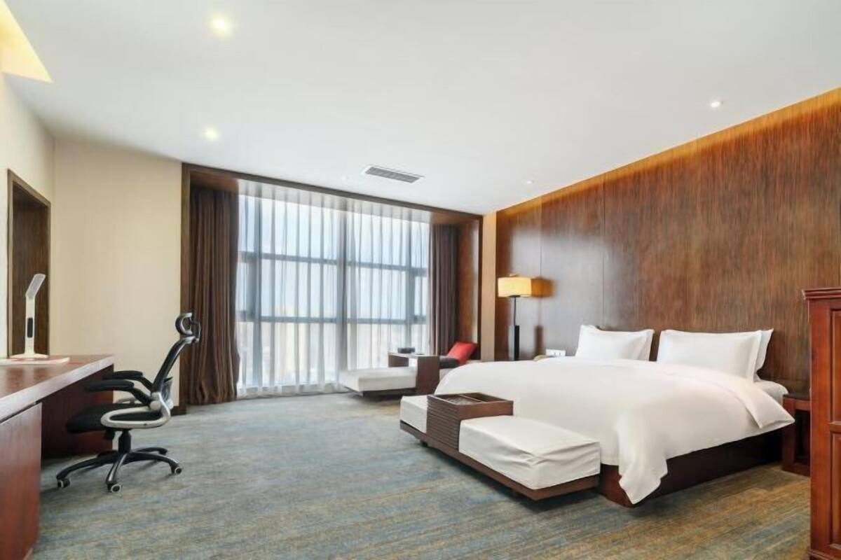 A hotel room with a large bed, a desk with a chair, a small table, and a window with curtains.