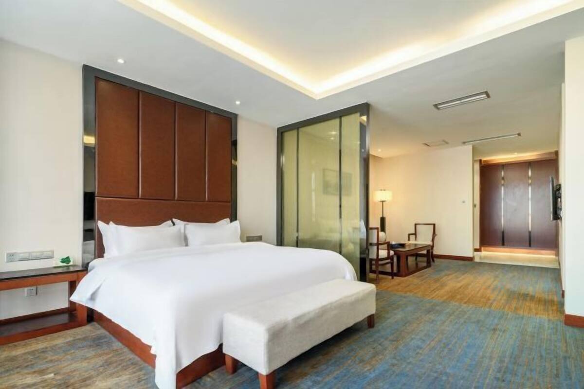 A hotel room with a large bed, a wooden headboard, a glass partition, a desk, and a chair.