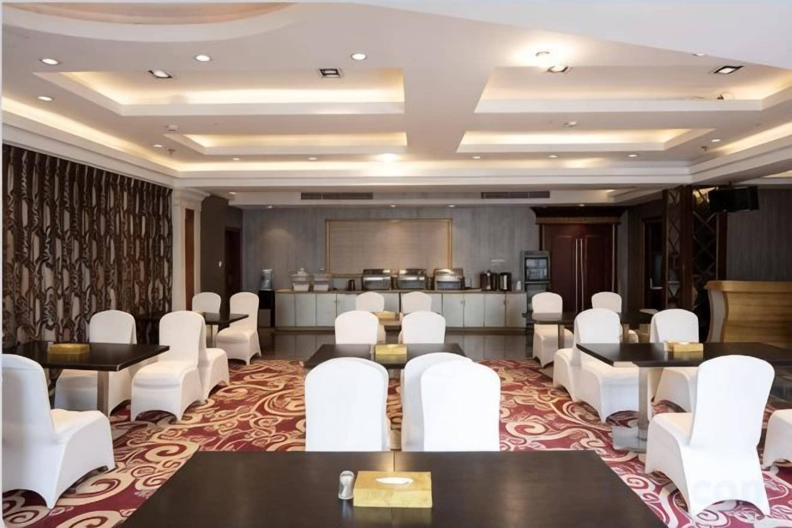 Un salón de conferencias con una mesa larga, sillas blancas, una alfombra estampada y una zona de cocina al fondo.