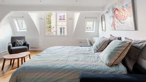 A spacious bedroom with a large bed, a chair, a desk, and a window with a view of buildings.