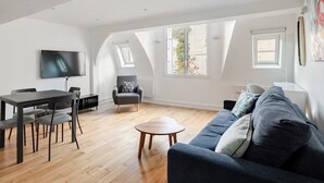 A spacious living area with a dark blue sofa, a dining table with chairs, a grey armchair, and a TV mounted on the wall.