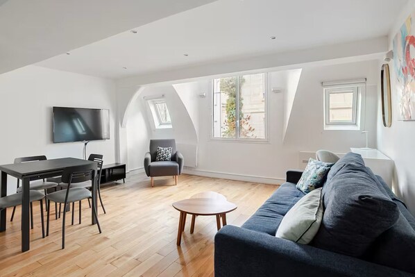 A spacious living area with a dark blue sofa, a dining table with chairs, a grey armchair, and a TV mounted on the wall.