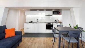 A modern kitchen with white cabinets, a microwave, and an oven. There is a dining table with chairs and a sofa with an orange pillow.