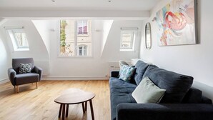 A spacious living room with a dark grey sofa, a wooden coffee table, and a black armchair with a patterned cushion. There are two windows providing natural light, and a colorful abstract painting on the wall.