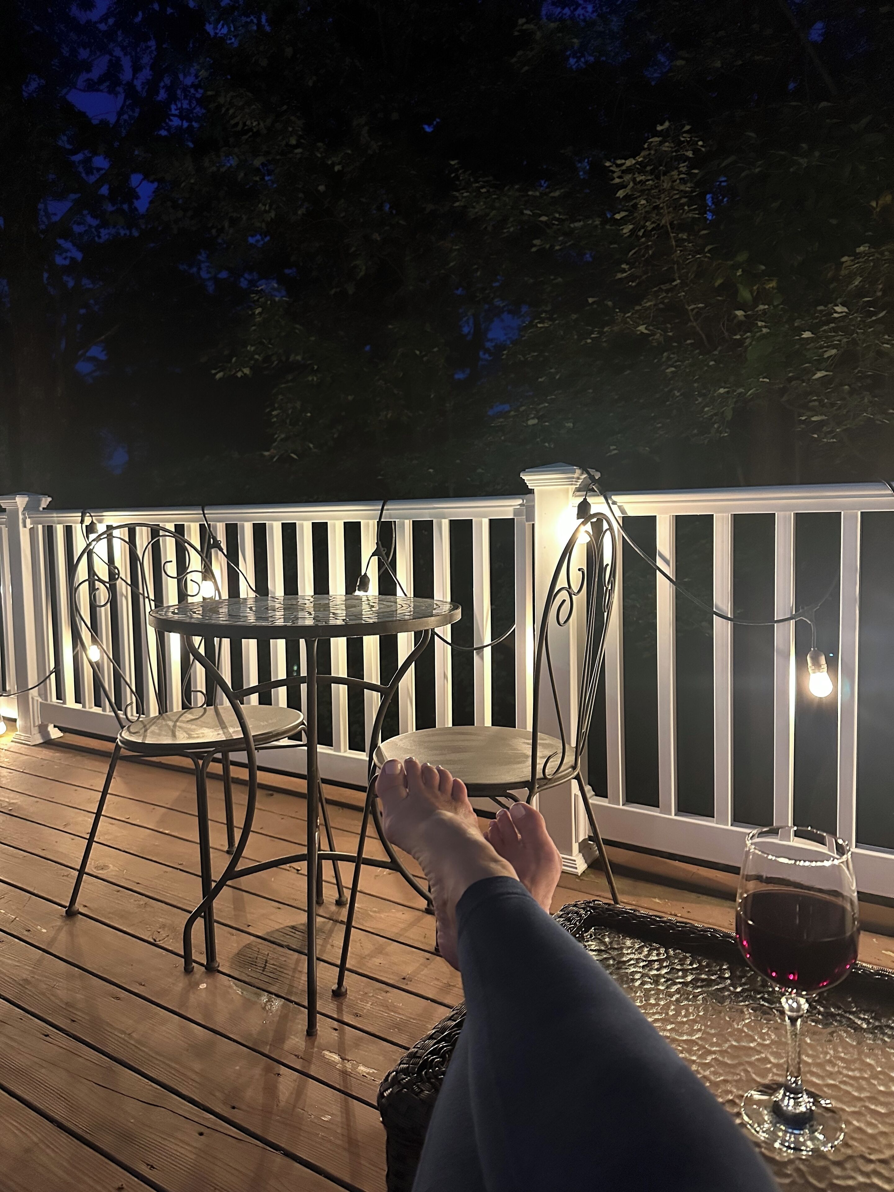 A wooden deck with a white railing, a round table, two chairs, and a glass of red wine.